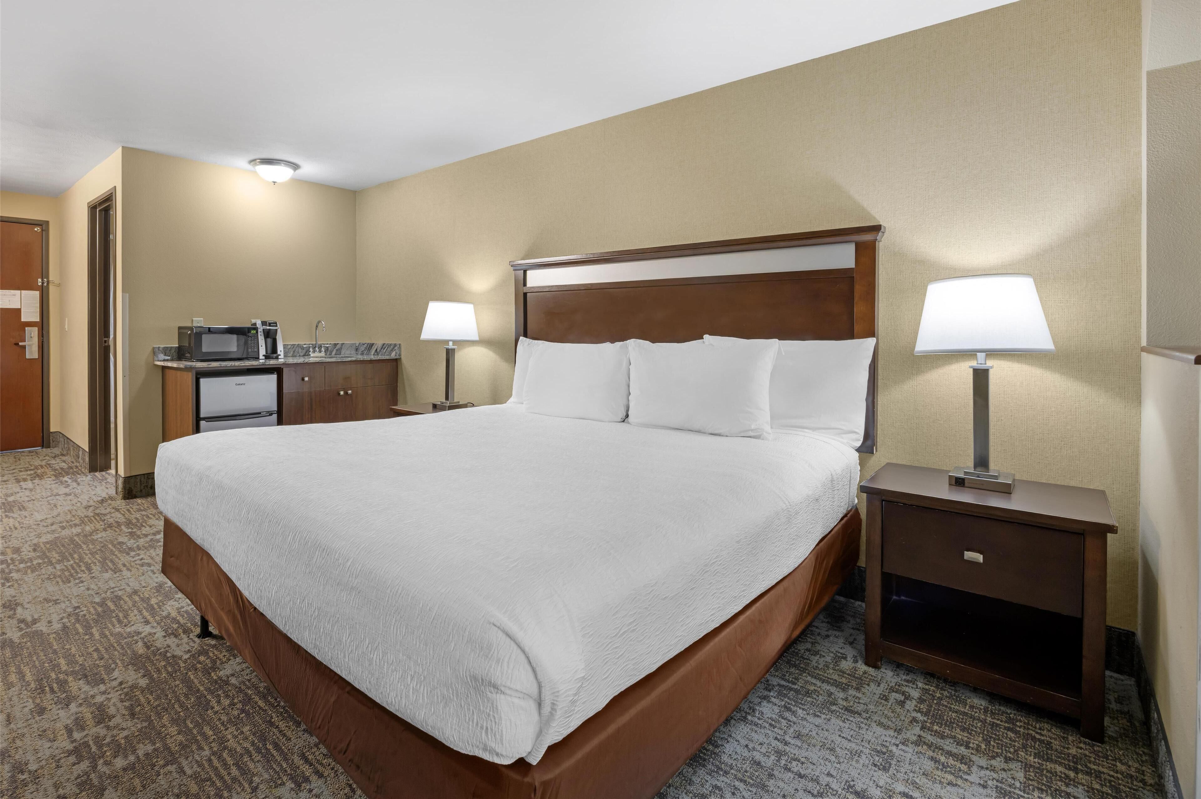 A hotel room with a large bed, two bedside tables with lamps, a small kitchenette, and a carpeted floor.