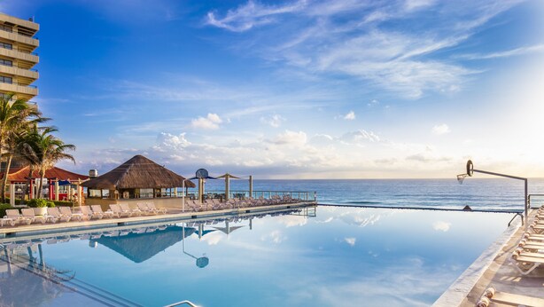 Piscina blu cristallina con lettini prendisole e una struttura a tetto di paglia, con vista su una spiaggia.