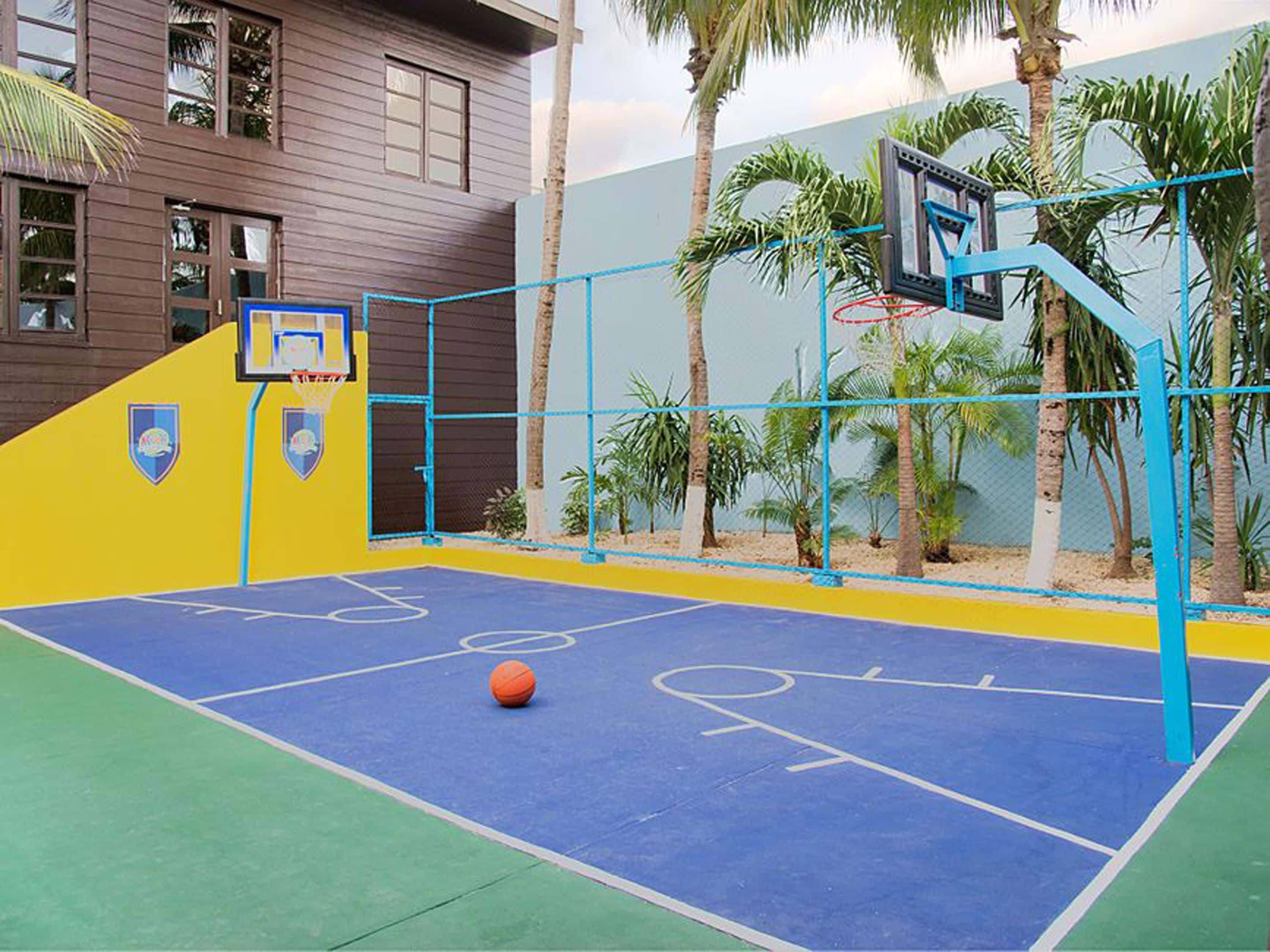 Una cancha de baloncesto al aire libre con superficie azul y amarilla, un aro de baloncesto y palmeras al fondo.