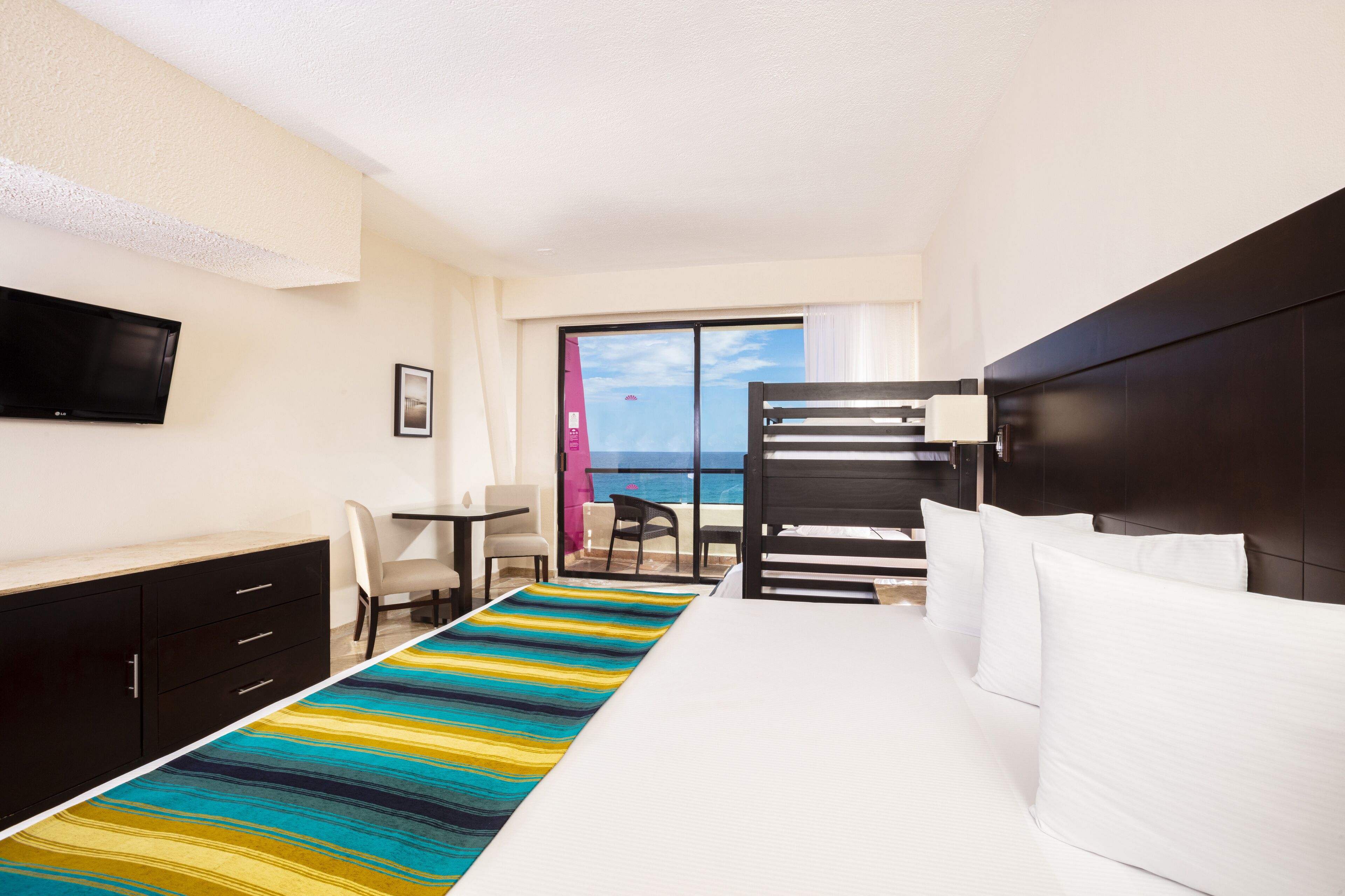 A hotel room with a bed, a desk, a chair, a TV, and a view of the ocean through a sliding door.