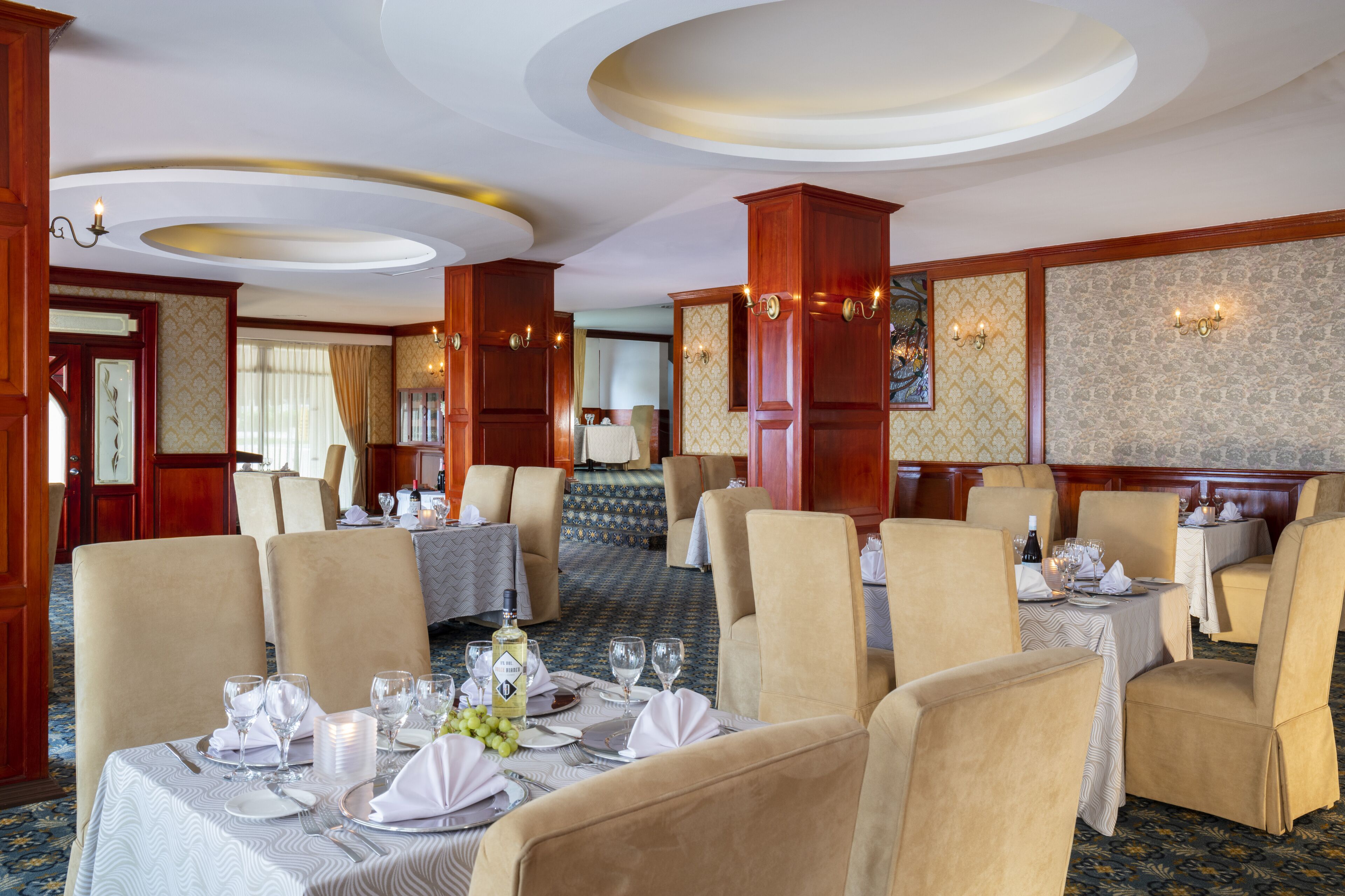 Un comedor de hotel con mesas dispuestas para el servicio, paneles de madera y una alfombra estampada.