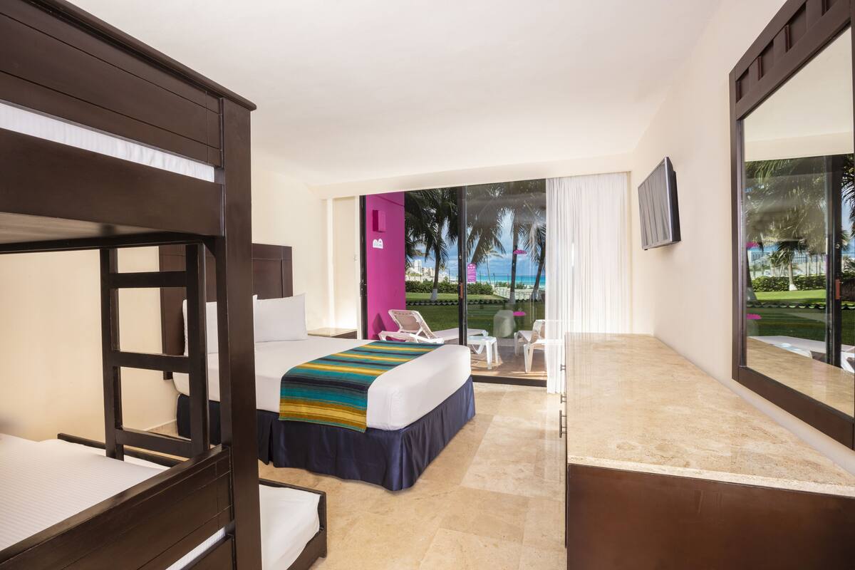 Una camera d'albergo con un letto a castello, un letto matrimoniale con una coperta colorata, un balcone con vista su palme e sull'oceano, e un grande specchio a parete.