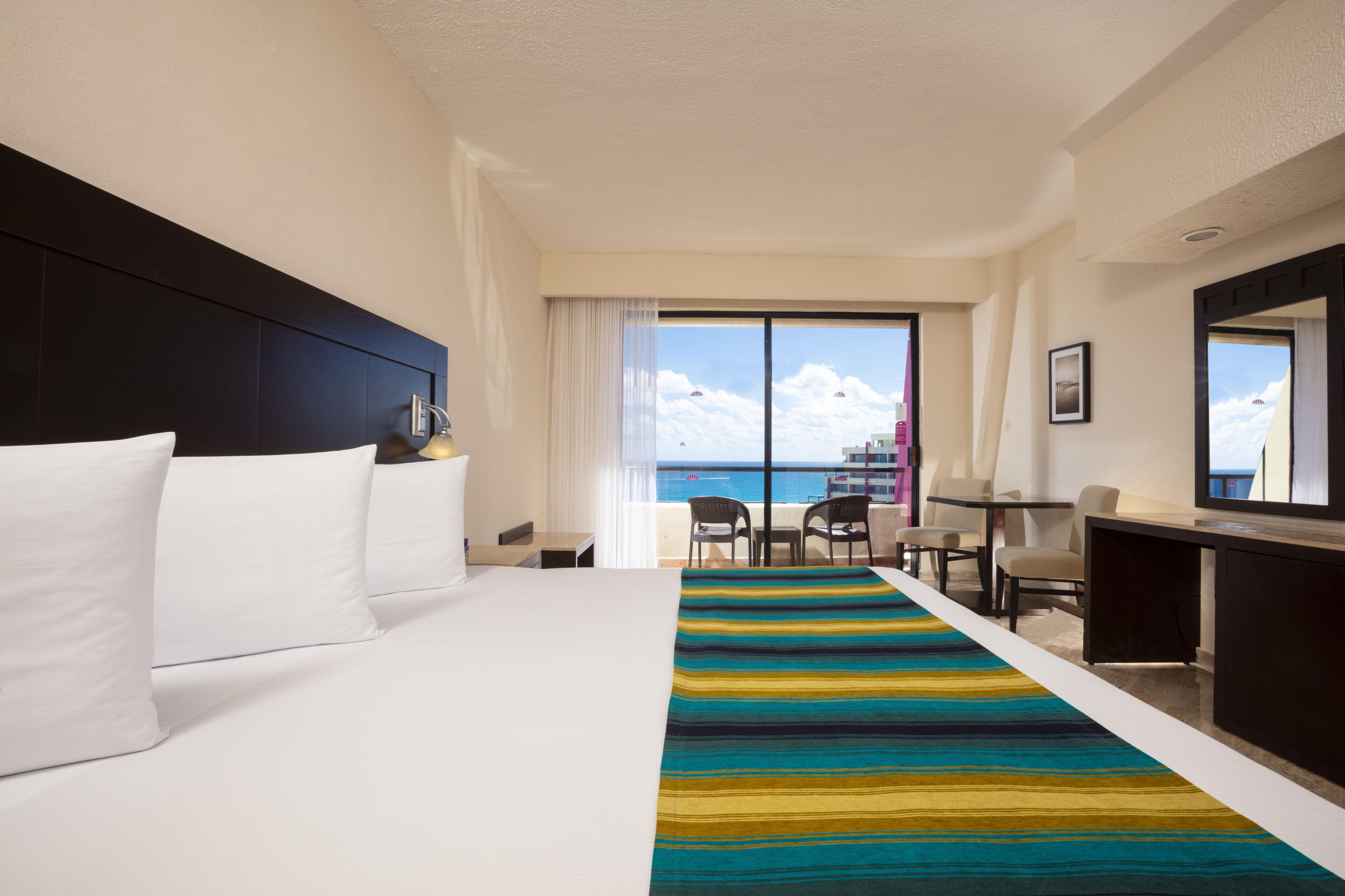 Une chambre d’hôtel avec un lit, un bureau, une chaise, une vue sur l’océan et un balcon équipé de chaises.