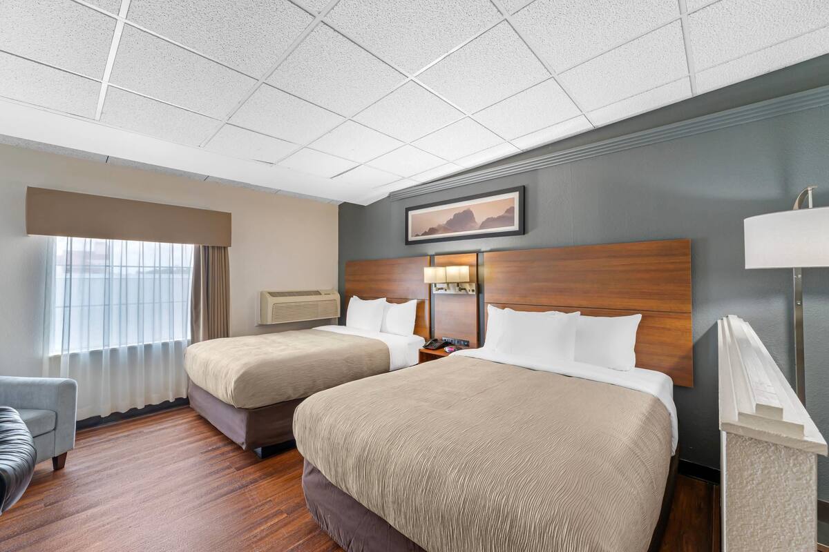 Habitación de hotel con dos camas, un escritorio, una silla, una ventana con cortinas y un cuadro en la pared.