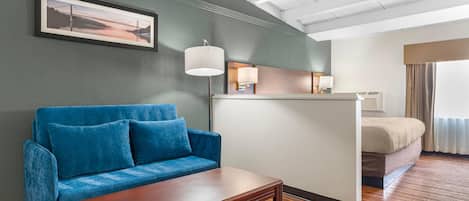 A hotel room with a blue sofa, a wooden coffee table, a bed with a beige comforter, a nightstand with a lamp, and a framed picture on the wall.
