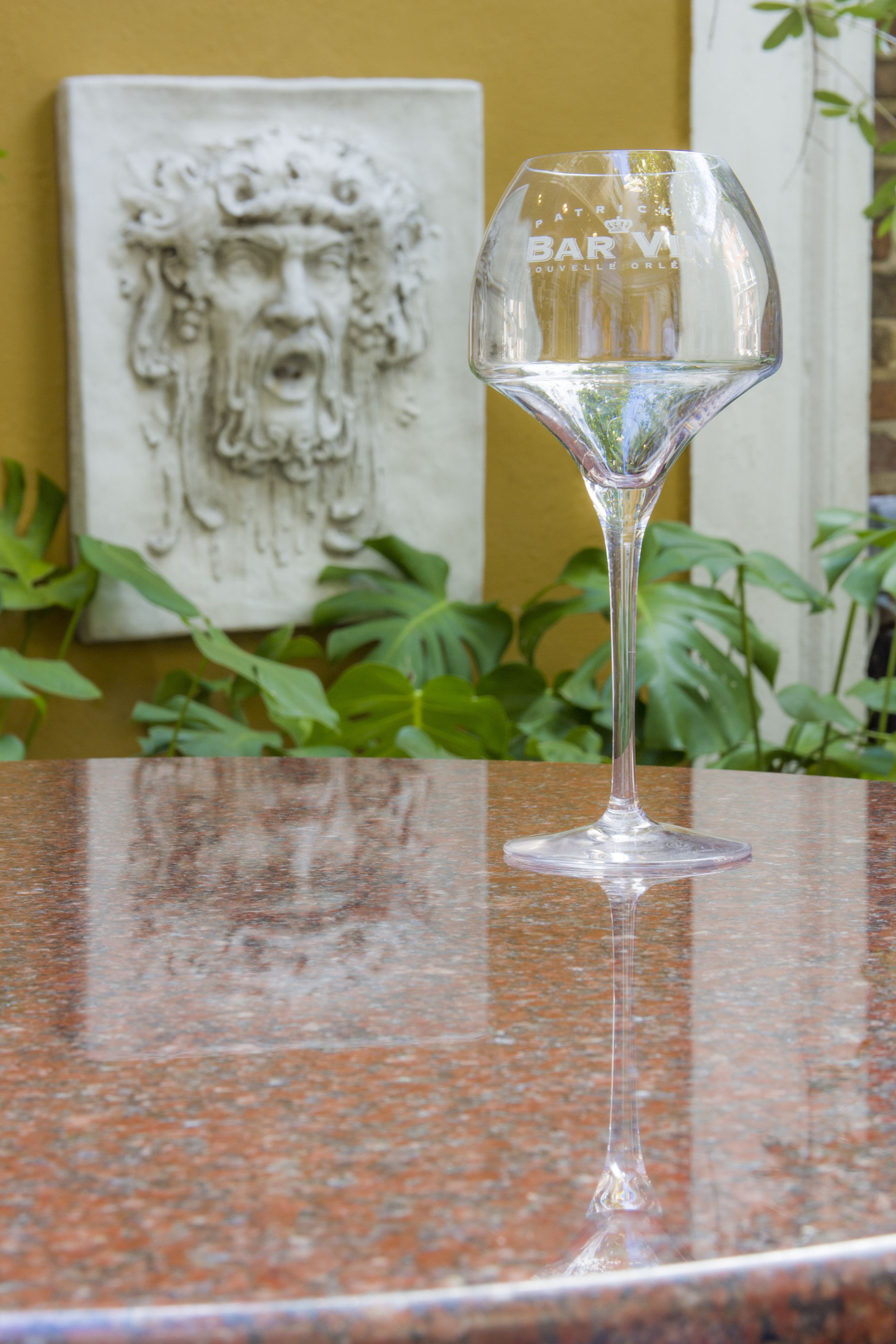 Taça de vinho de vidro transparente sobre bancada de granito, com uma escultura e plantas verdes ao fundo.