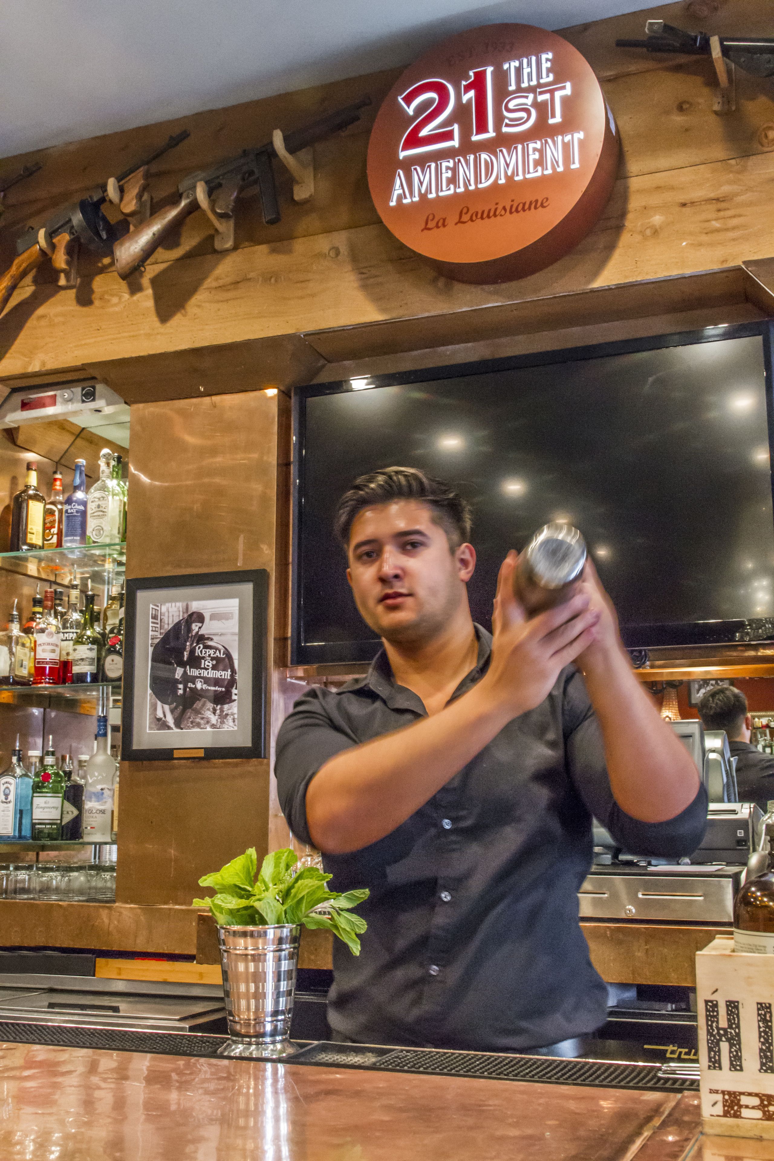 Ein Barkeeper bereitet ein Getränk an der Bar zu, wo ein Schild mit der Aufschrift "21st Amendment" und ein gerahmtes Bild einer historischen Persönlichkeit zu sehen sind.