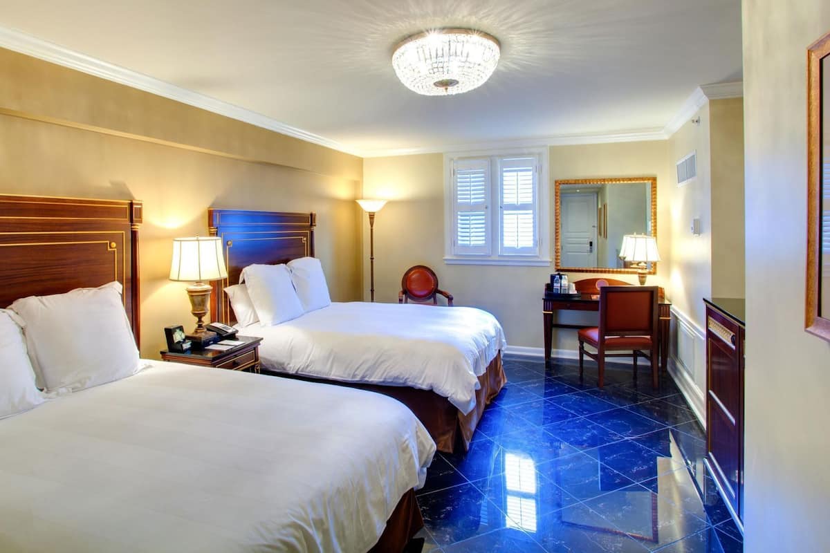 Une chambre d’hôtel avec deux lits, un bureau, une chaise, une lampe, un grand miroir et un lustre.
