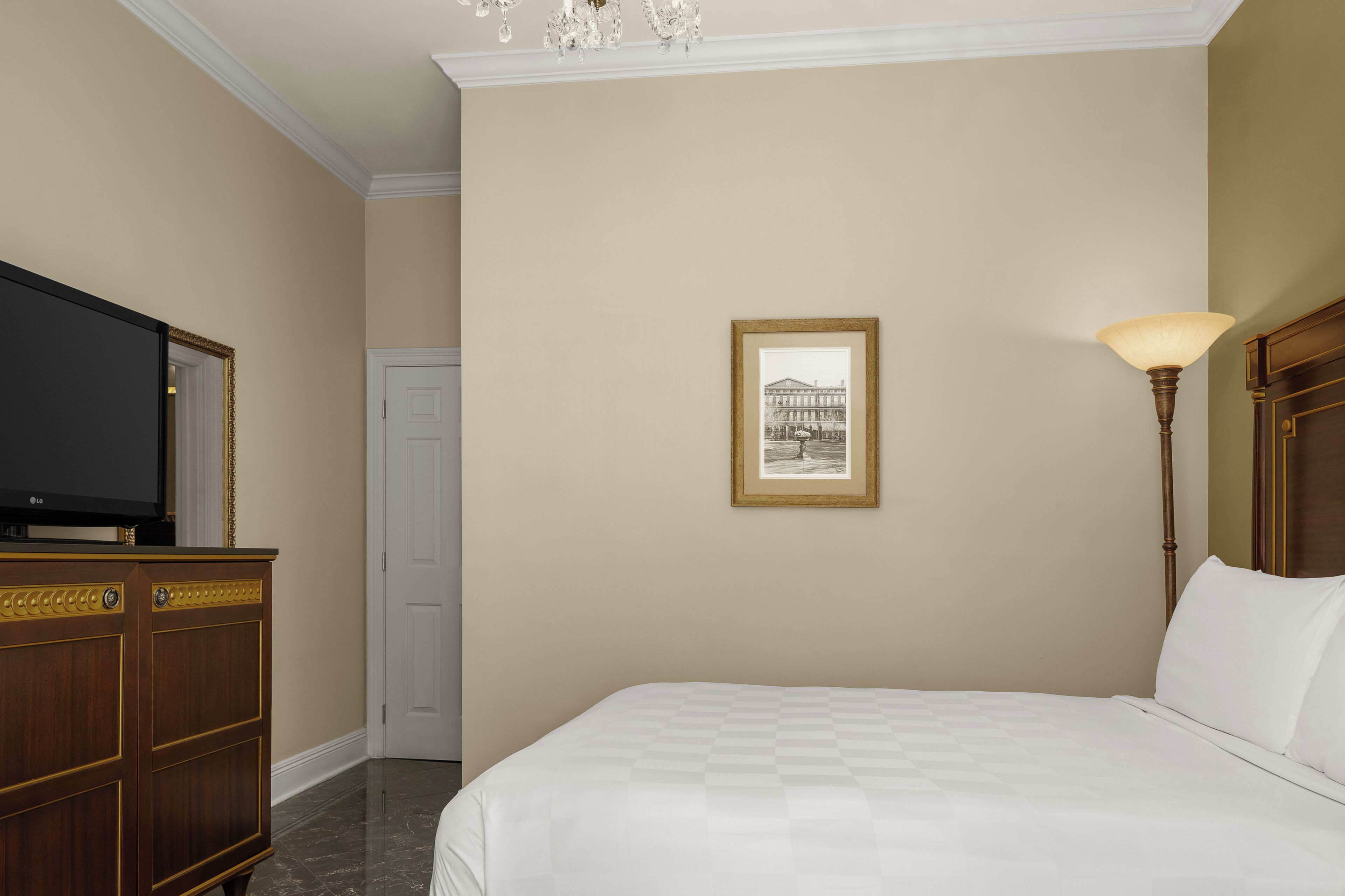 A hotel room with a large bed, a television on a wooden cabinet, a framed picture on the wall, and a floor lamp.