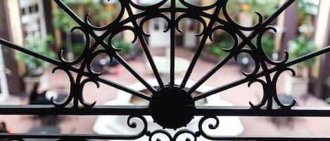 Un garde-corps de balcon en fer forgé, orné de motifs complexes, donnant sur une cour intérieure avec une fontaine et de la verdure.