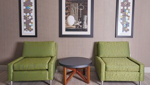 Two green armchairs with a geometric pattern, a round table with wooden legs, and three framed artworks on the wall.