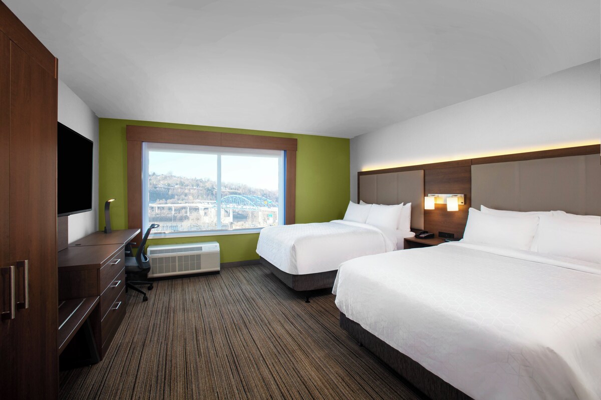 Une chambre d’hôtel avec deux lits, un bureau, une chaise, un téléviseur à écran plat, une fenêtre donnant sur la ville et un radiateur fixé au mur.