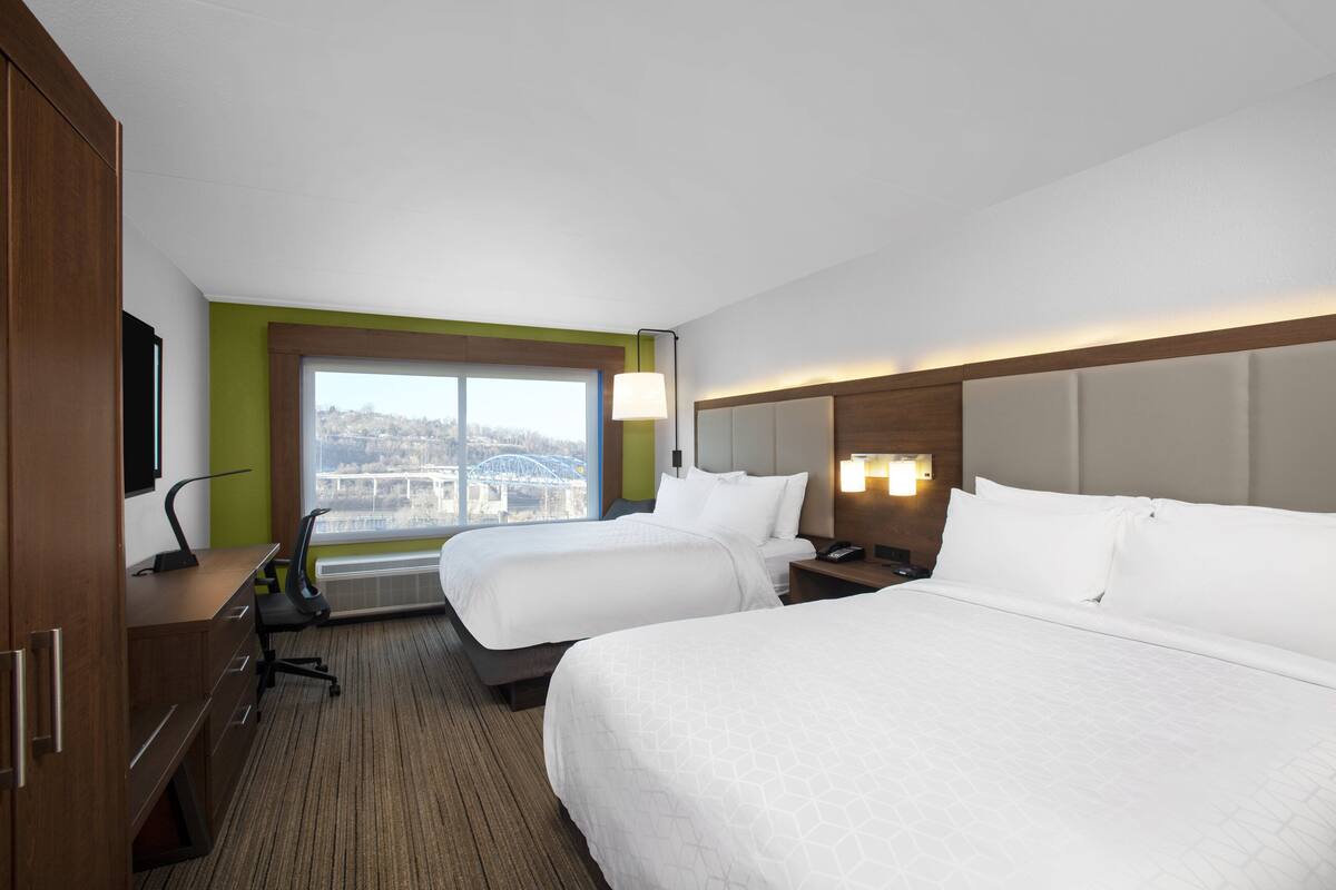 A hotel room with two beds, a desk, a chair, a TV, a window with a city view, and a lamp.
