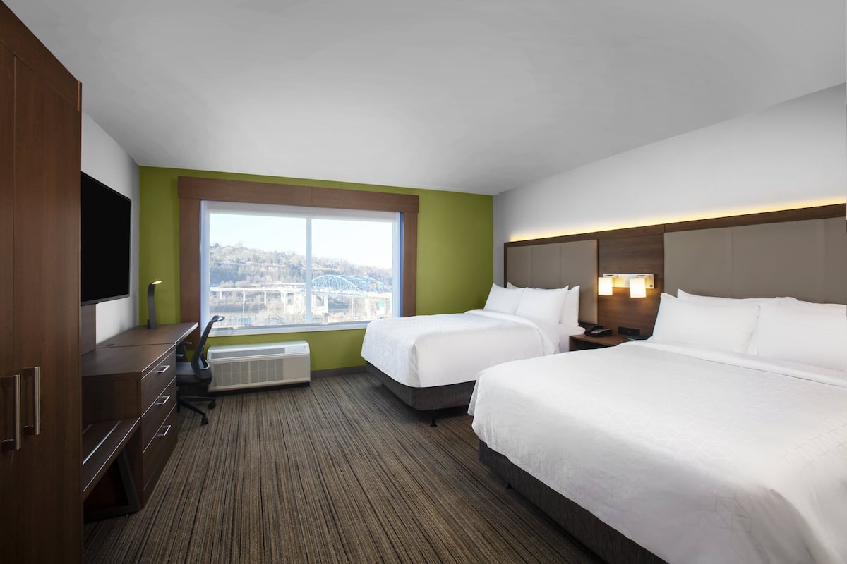 A hotel room with two beds, a desk, a chair, a flat-screen TV, a window with a city view, and a wall-mounted heater.