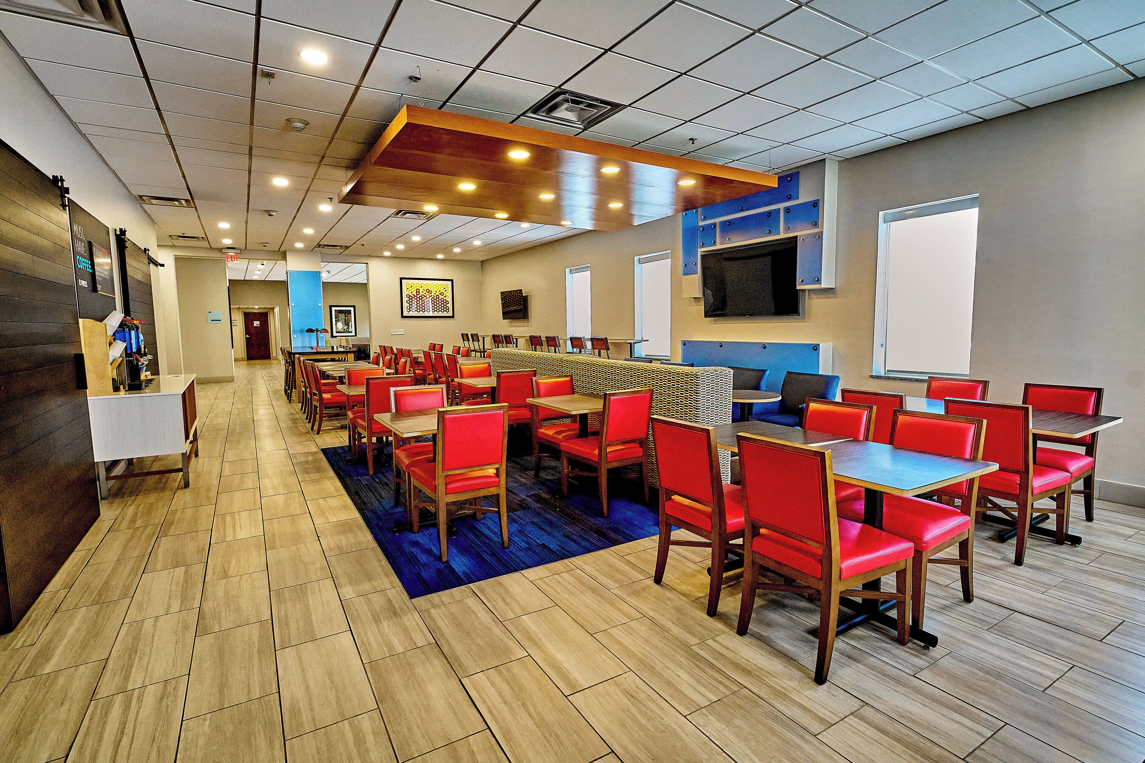 A hotel dining area with multiple tables and chairs, a blue carpet, and a television mounted on the wall.