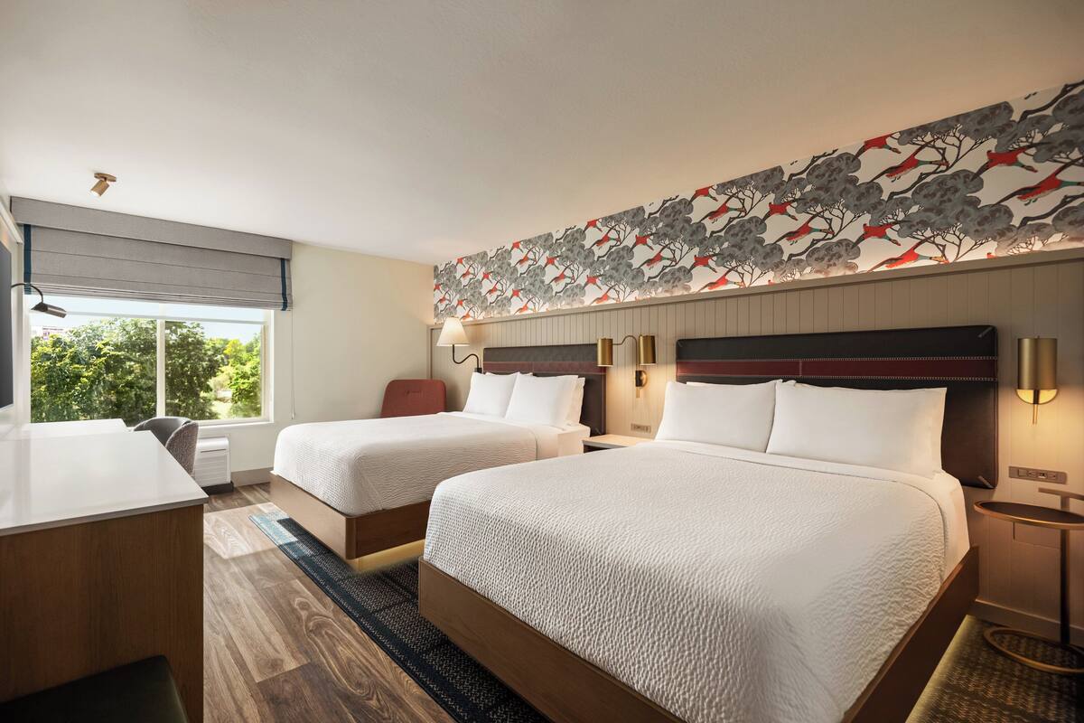 A hotel room with two beds, a desk, a chair, a window with a view of trees, and a decorative wall with a tree and bird pattern.