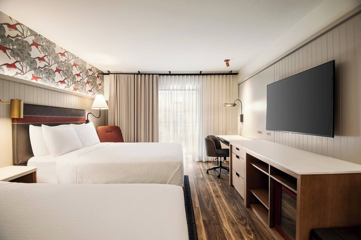 A hotel room with two beds, a desk, a chair, a TV, and a large window with curtains.