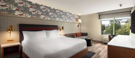 A hotel room with a large bed, a desk, a chair, a sofa, a lamp, and a window with a view.
