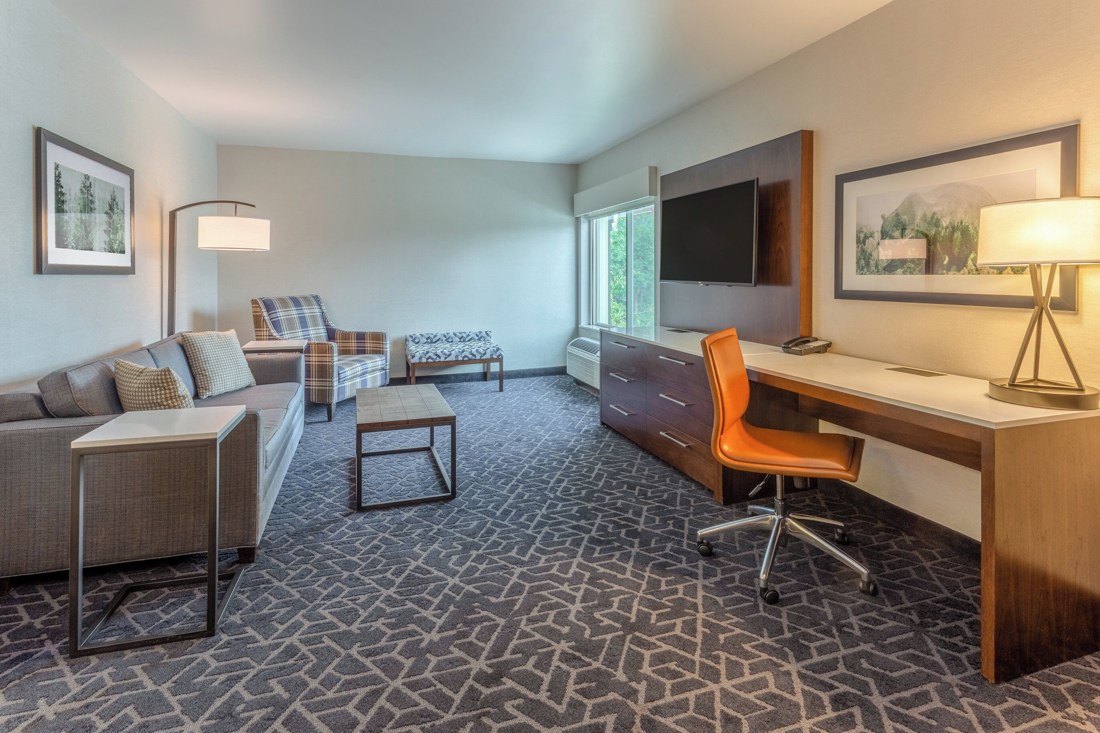 A hotel room with a sofa, a small table, a desk with an orange chair, a TV, and a lamp.