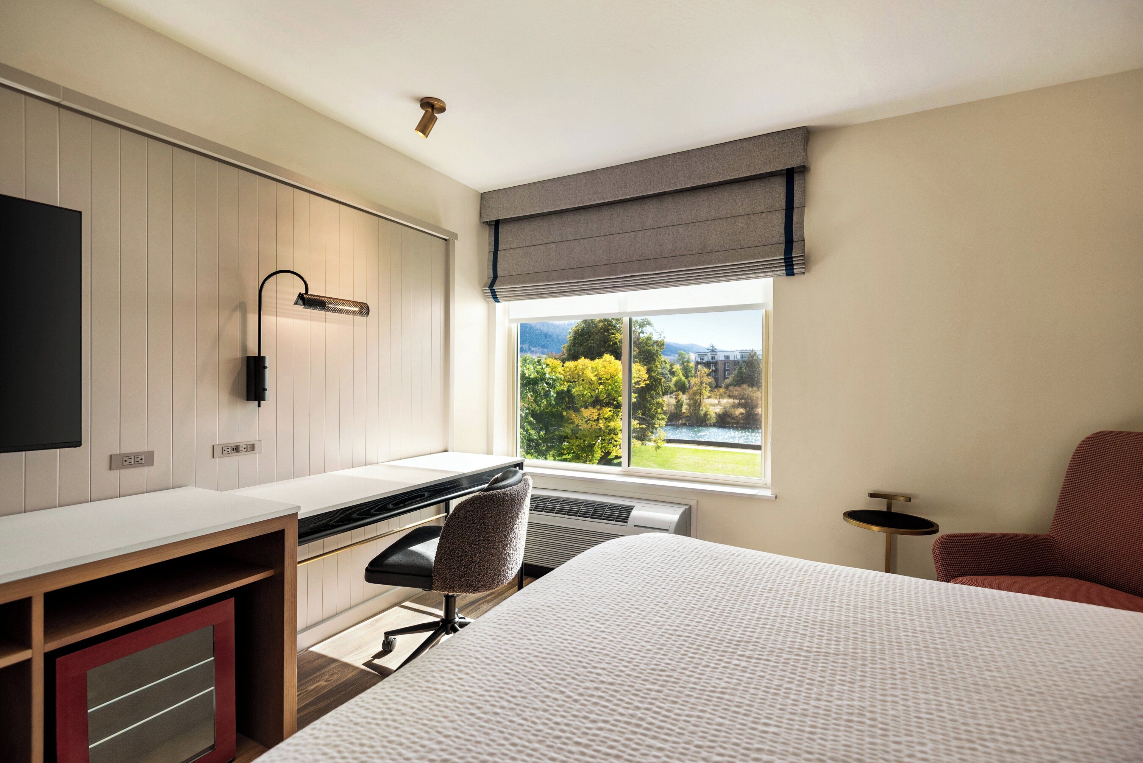 A hotel room with a bed, desk, chair, and a window with a view of greenery and mountains.