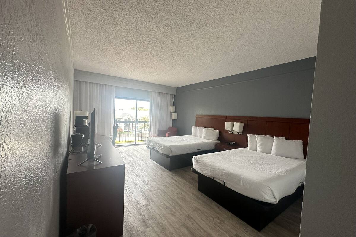 A hotel room with two beds, a desk with a computer, a TV, and a balcony with a view.