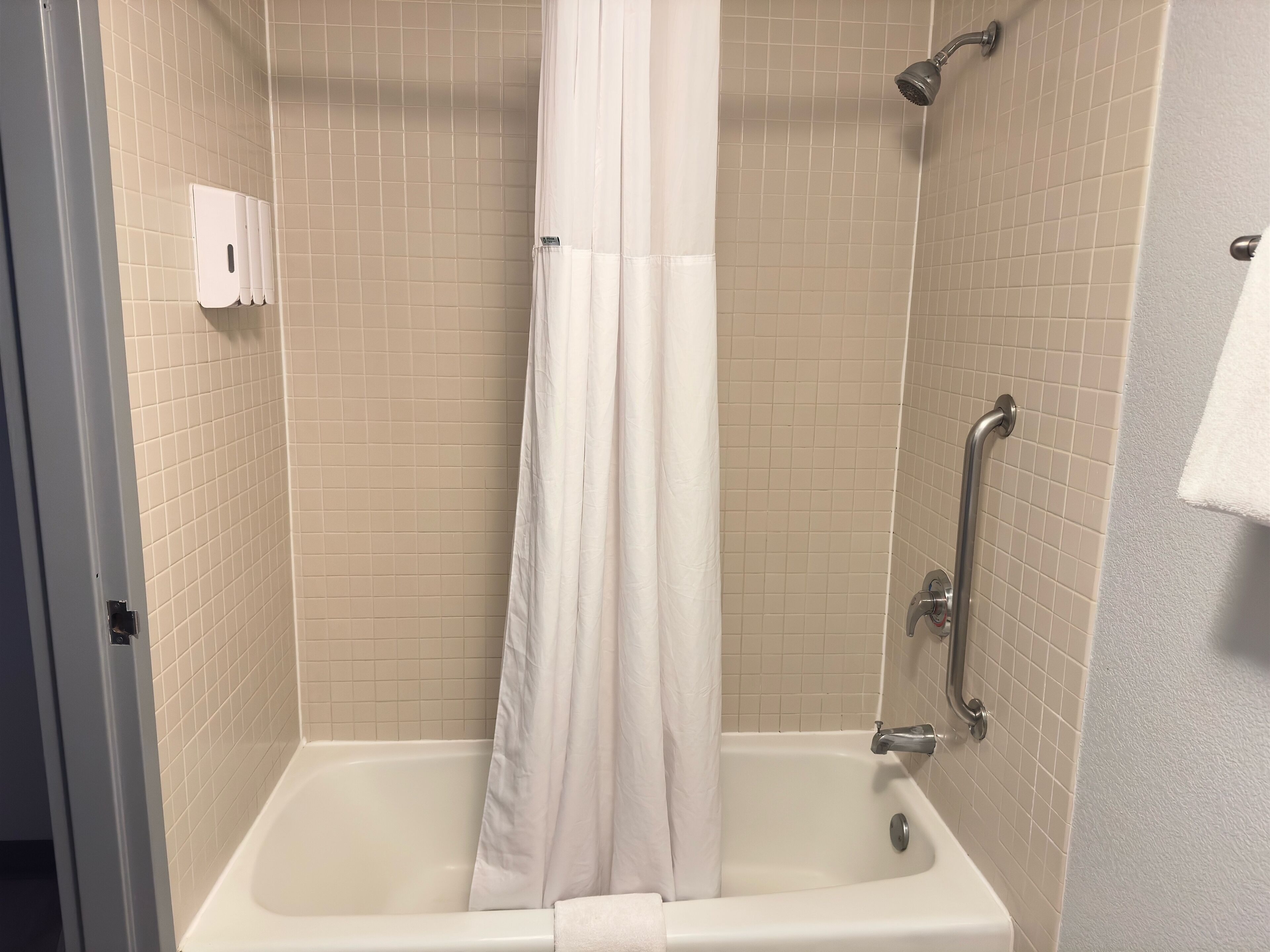 Ducha con cortina blanca, paredes de azulejos color beige y un cabezal de ducha de mano.