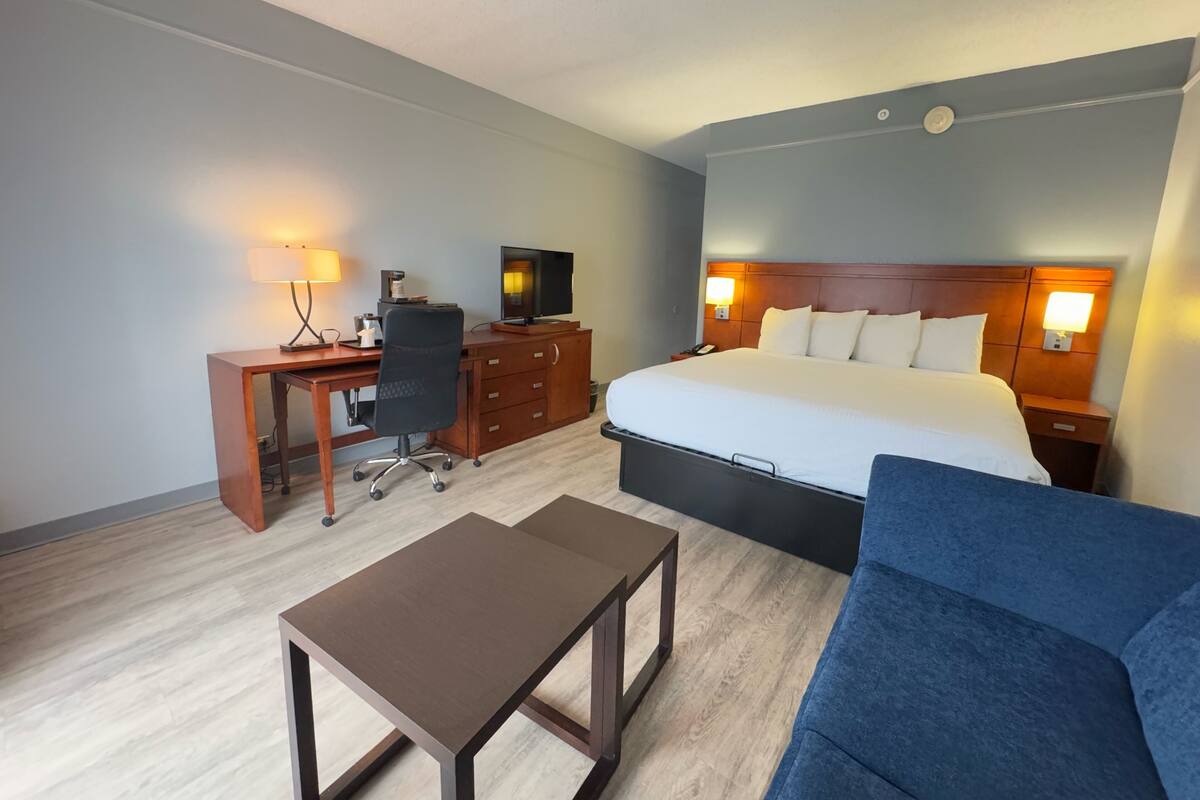 A hotel room with a bed, a desk with a chair, a television, a coffee maker, and a nightstand with a lamp.