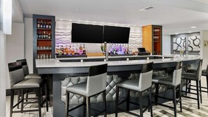 A modern bar area with a large flat-screen TV, bar stools, and shelves stocked with bottles and glasses.
