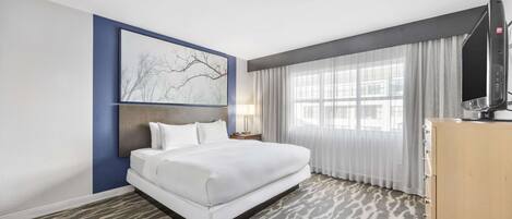 A hotel room with a large bed, a flat-screen TV, a desk with a lamp, and a window with sheer curtains.