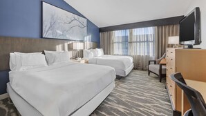 A hotel room with two beds, a desk, a television, and a large window with curtains.