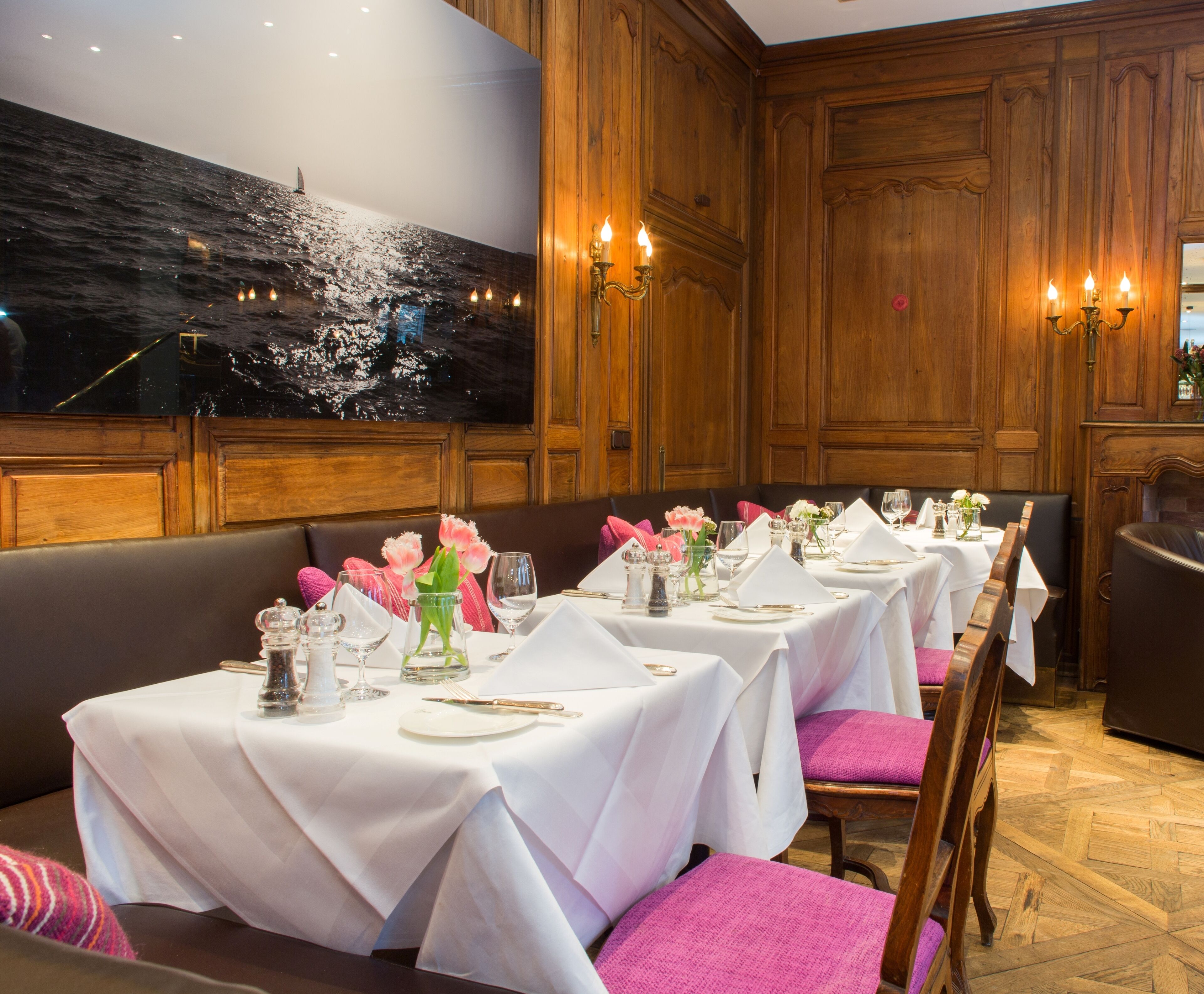 A dining area with tables set for service, wooden paneling, and a large painting of a seascape on the wall.