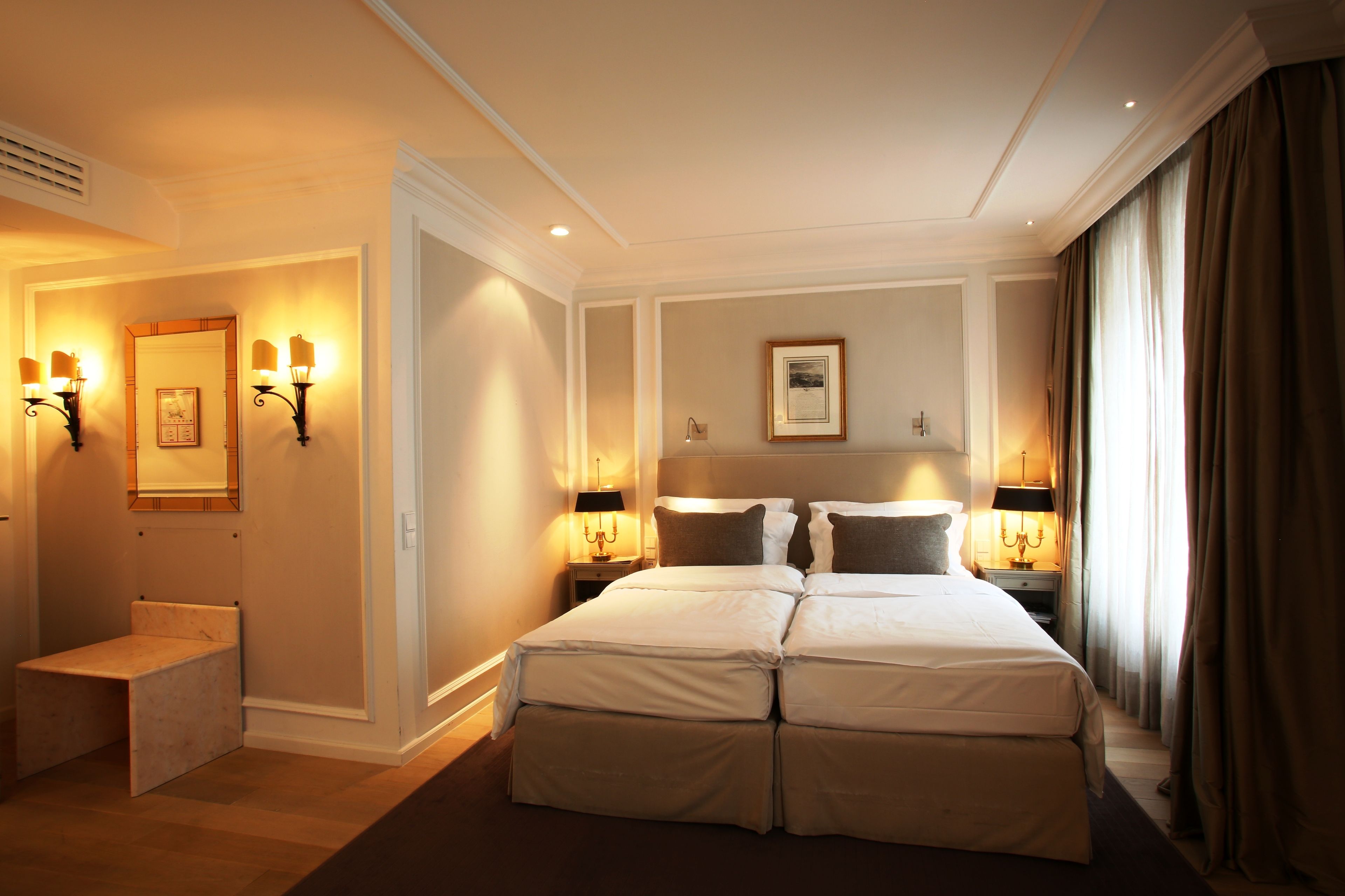 Una habitación de hotel con una cama grande, lámparas de noche, un pequeño banco y cuadros en las paredes.