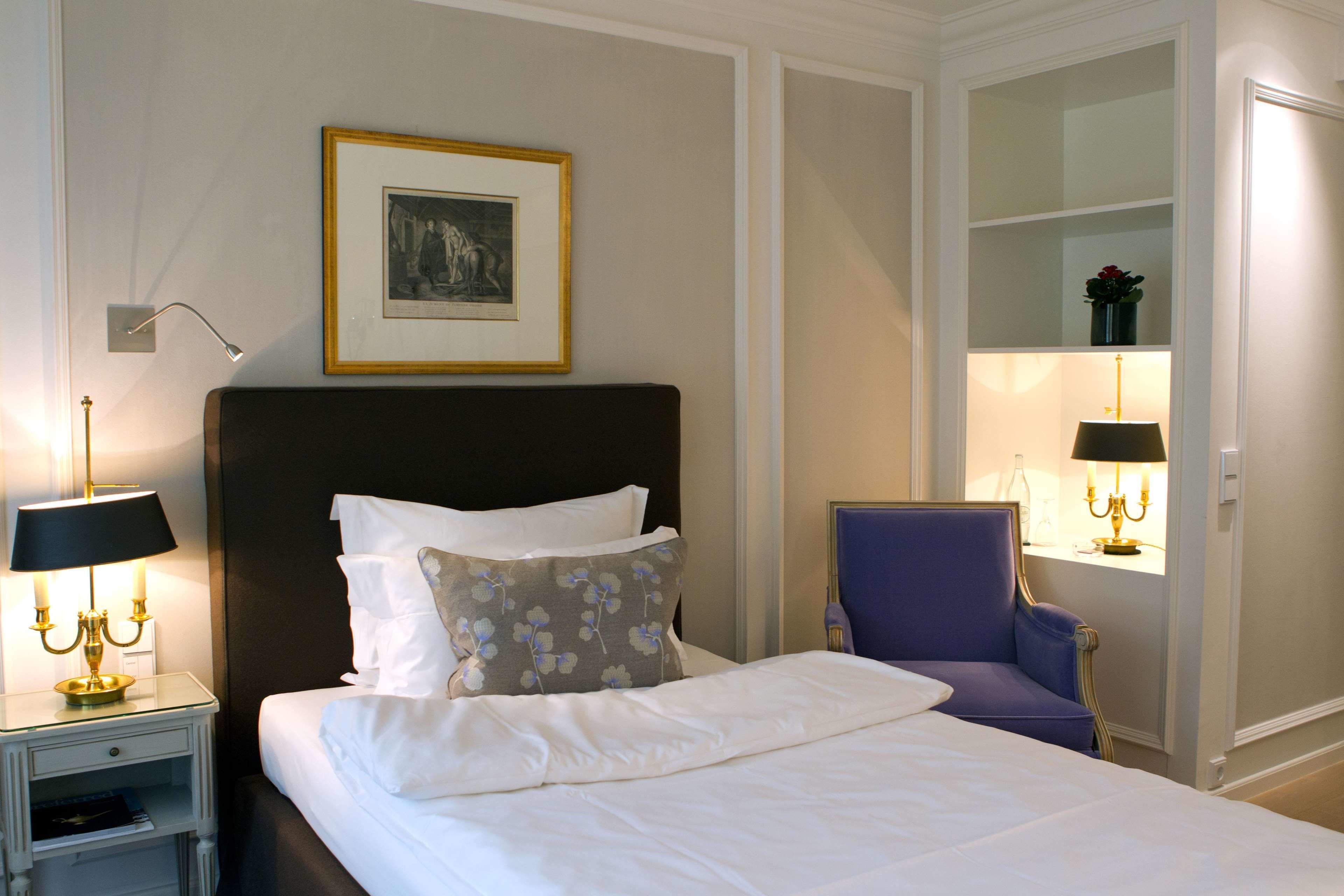 A neatly made bed with white linens and a dark headboard, flanked by two bedside tables with lamps. A framed picture hangs above the bed, and a chair is positioned next to the bed.