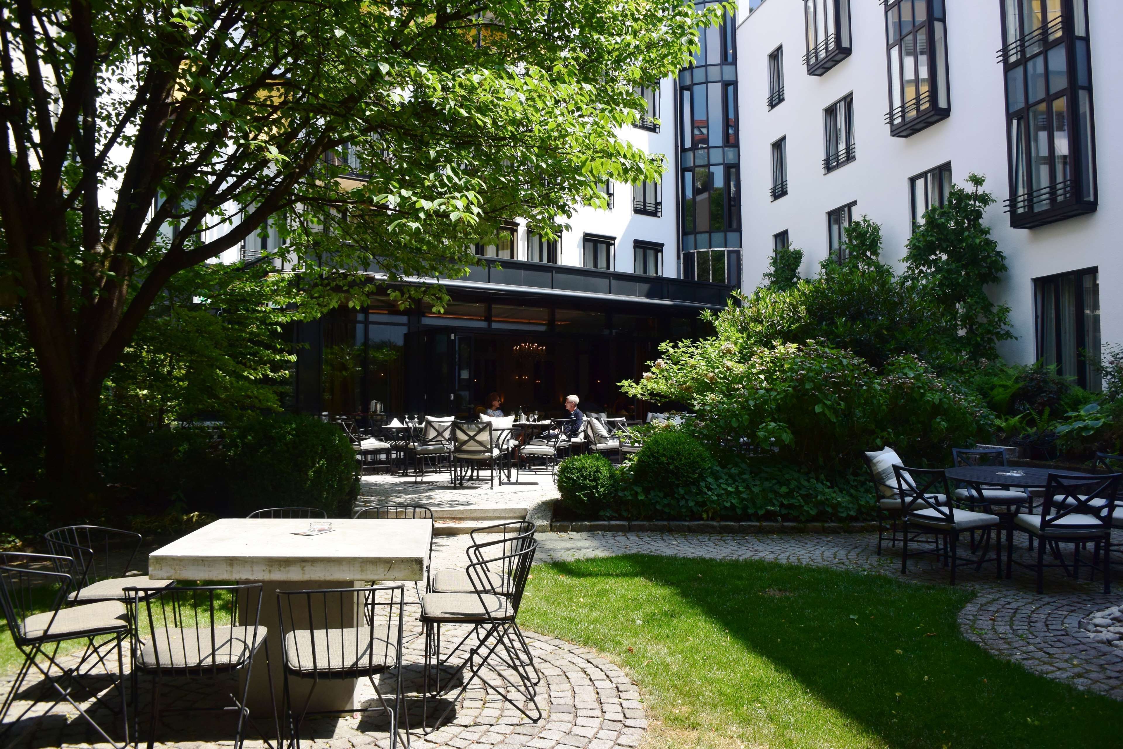 An outdoor patio area with tables and chairs, surrounded by greenery and a building with large windows.