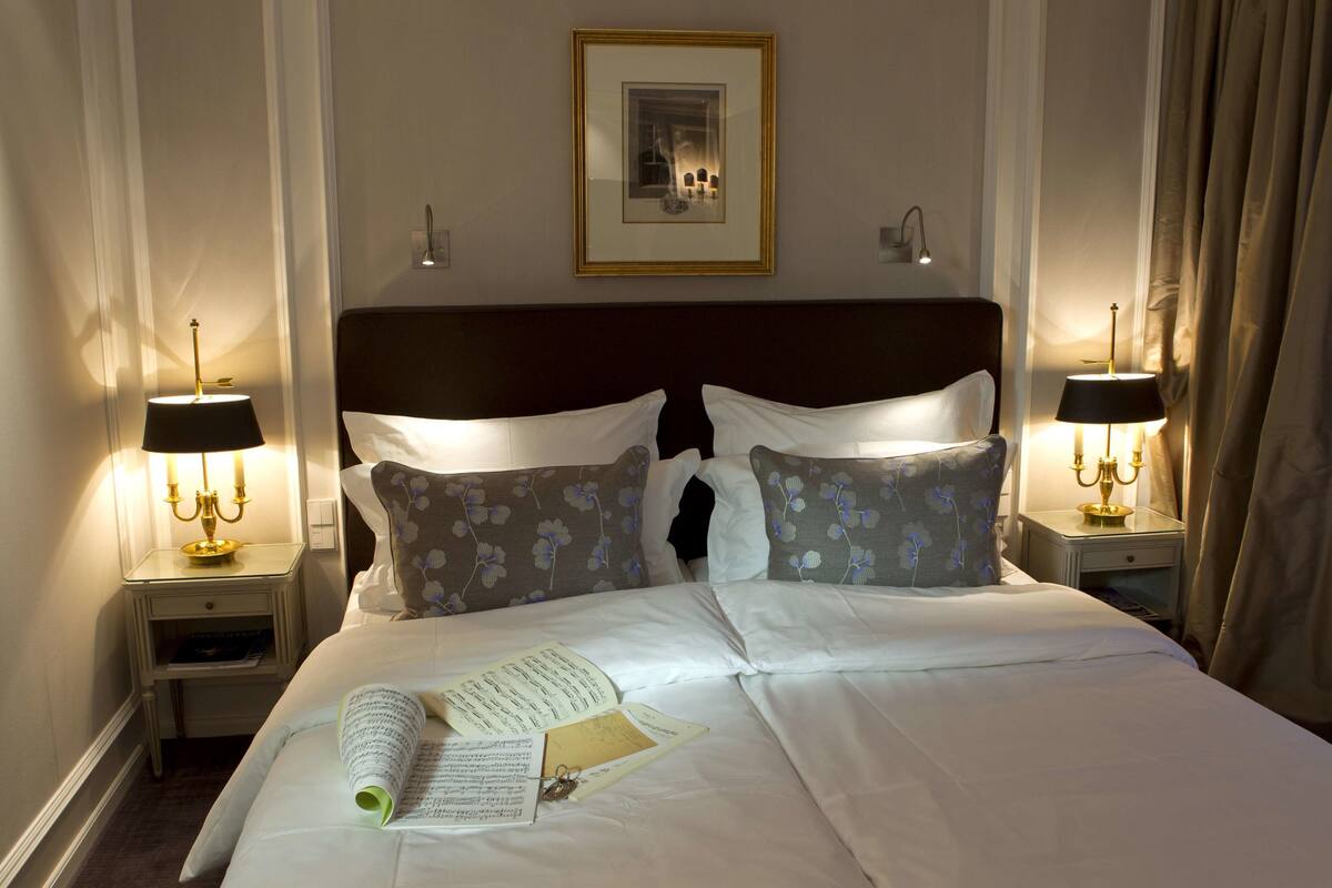Una cama bien hecha con almohadones florales, un cuadro enmarcado encima y dos mesitas de noche con lámparas.