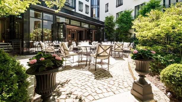 Un patio al aire libre con mesas y sillas, rodeado de vegetación y un edificio con grandes ventanales.