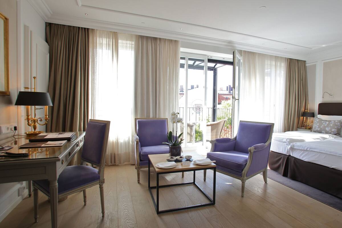 Una habitación de hotel con cama, dos sillones, una mesita, un escritorio y una ventana con cortinas.