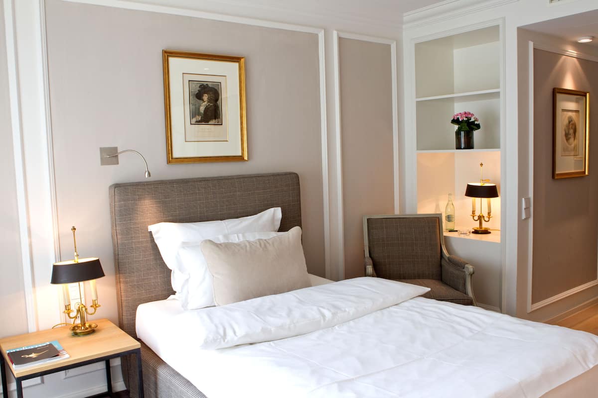 Una cama bien hecha con sábanas blancas, cabecera y mesita de noche con lámpara. Encima de la cama hay un cuadro enmarcado en la pared, y a la derecha, un estante pequeño con un jarrón de flores.