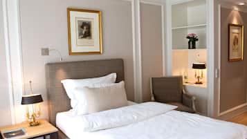 Una cama bien hecha con sábanas blancas, cabecera y mesita de noche con lámpara. Encima de la cama hay un cuadro enmarcado en la pared, y a la derecha, un estante pequeño con un jarrón de flores.