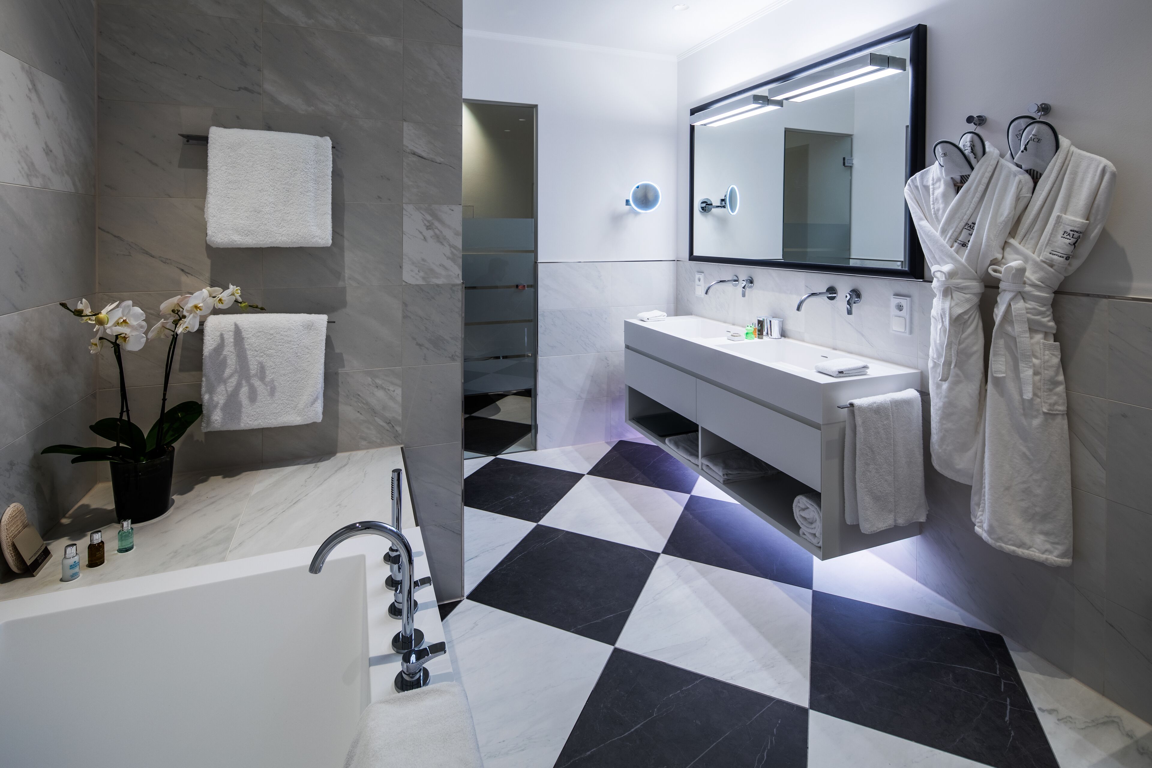 A modern bathroom with a checkered floor, a large mirror, a bathtub, and two white towels hanging on the wall.