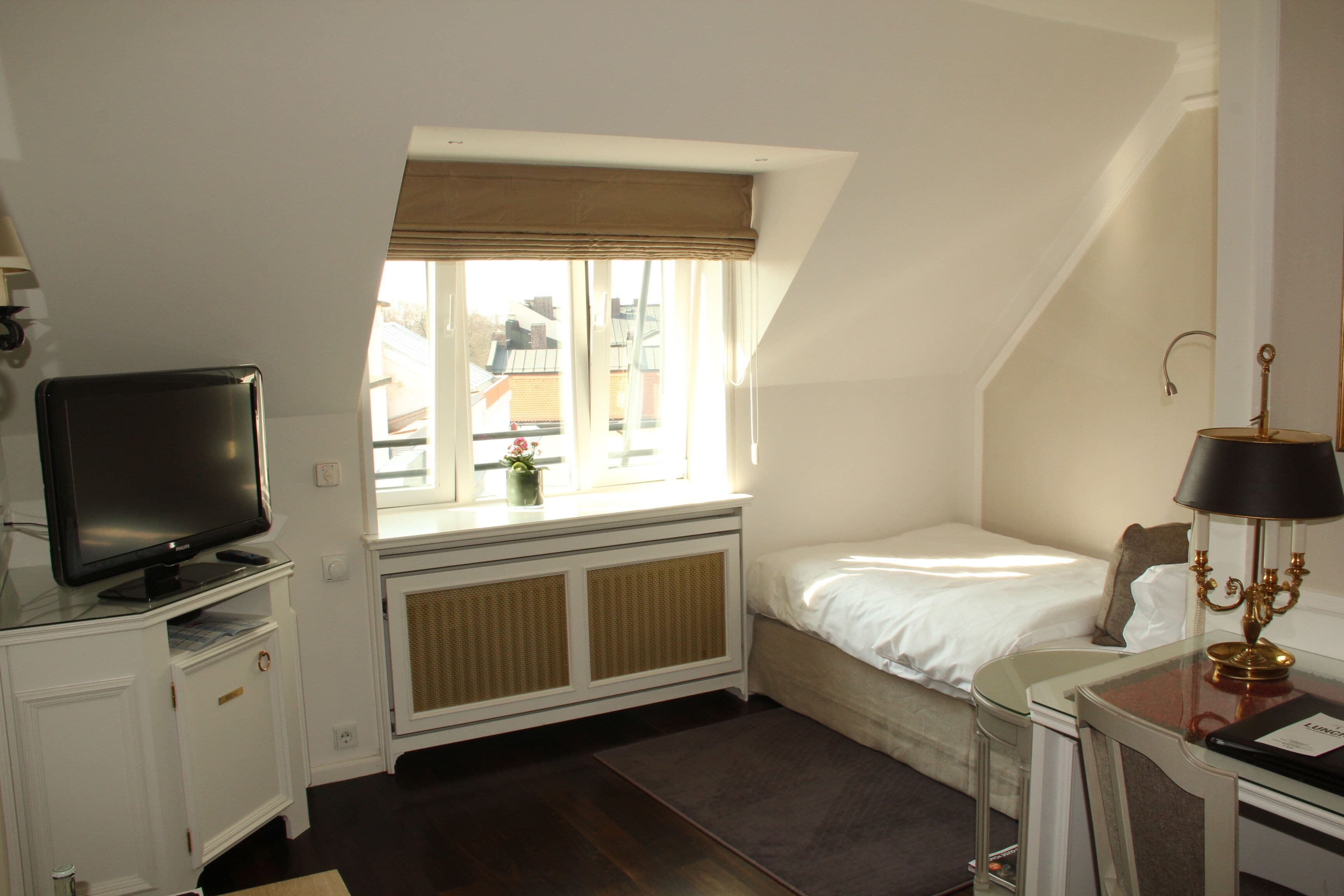 A hotel room with a bed, a TV, a window with a view of buildings, a radiator, and a lamp on a side table.