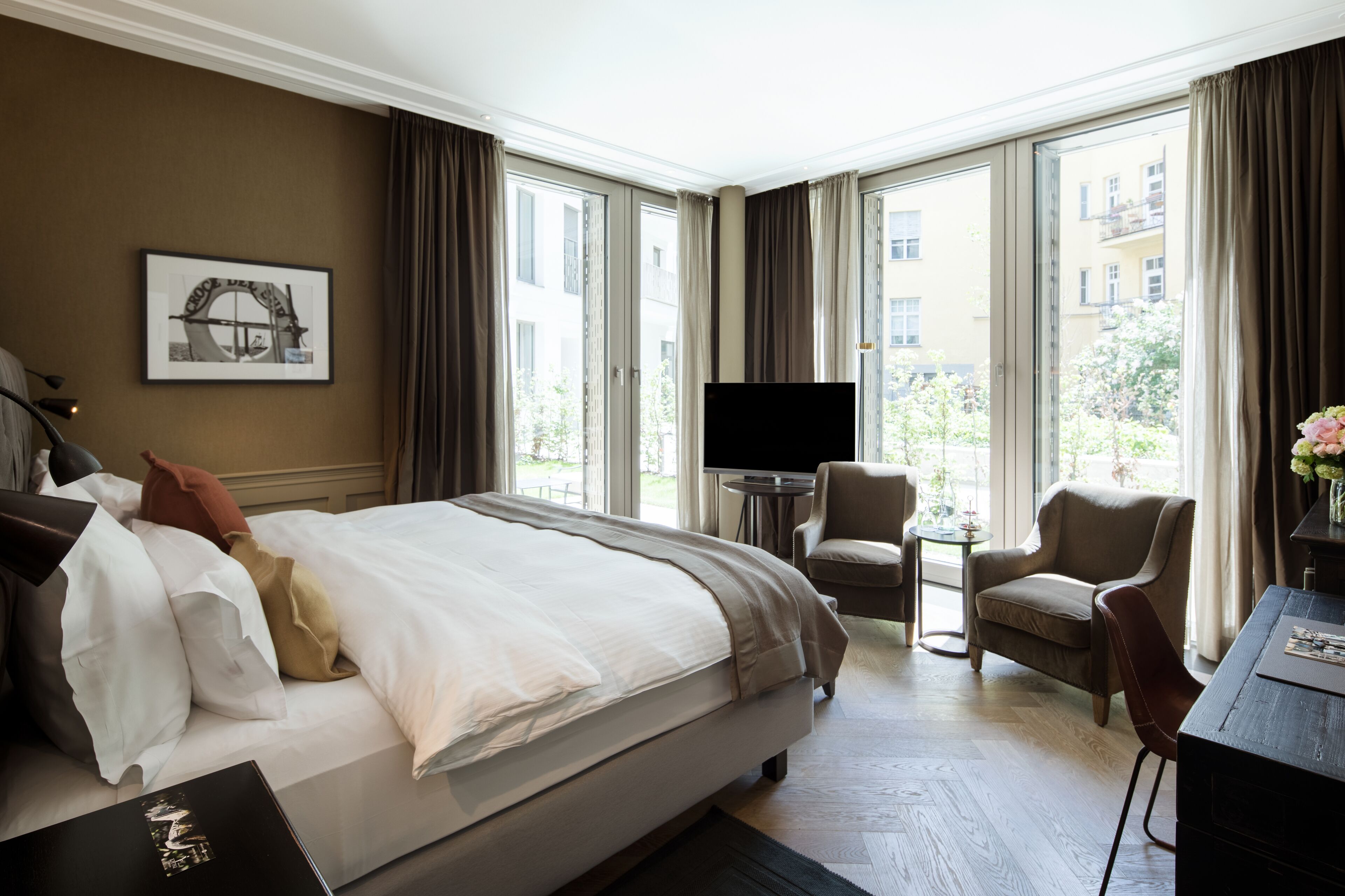 A hotel room with a large bed, a TV, a small table, and a chair. There is a window with curtains and a view of buildings outside.