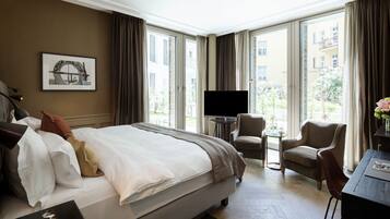 A hotel room with a large bed, a TV, a small table, and a chair. There is a window with curtains and a view of buildings outside.