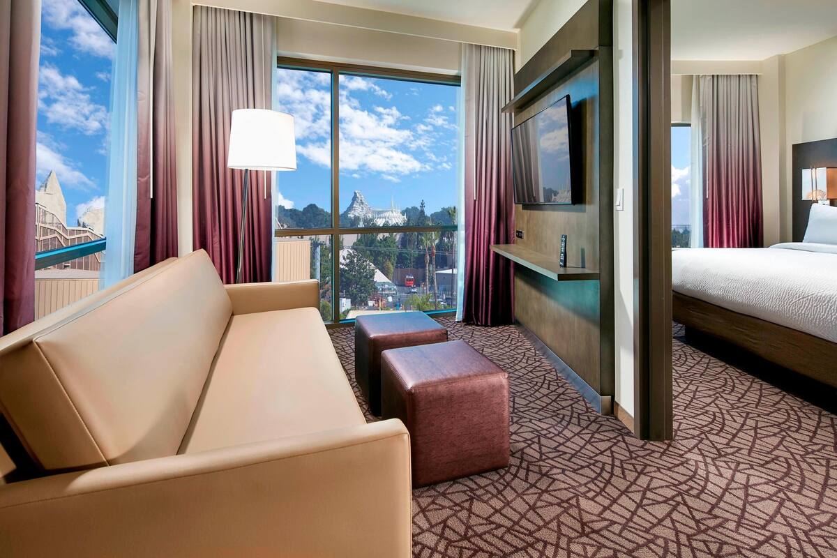 A hotel room with a large window, a sofa, a bed, a TV, and a patterned carpet.