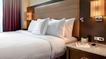 Una cama bien hecha con ropa blanca, una mesita de noche con una lámpara, una planta pequeña y un teléfono inteligente.