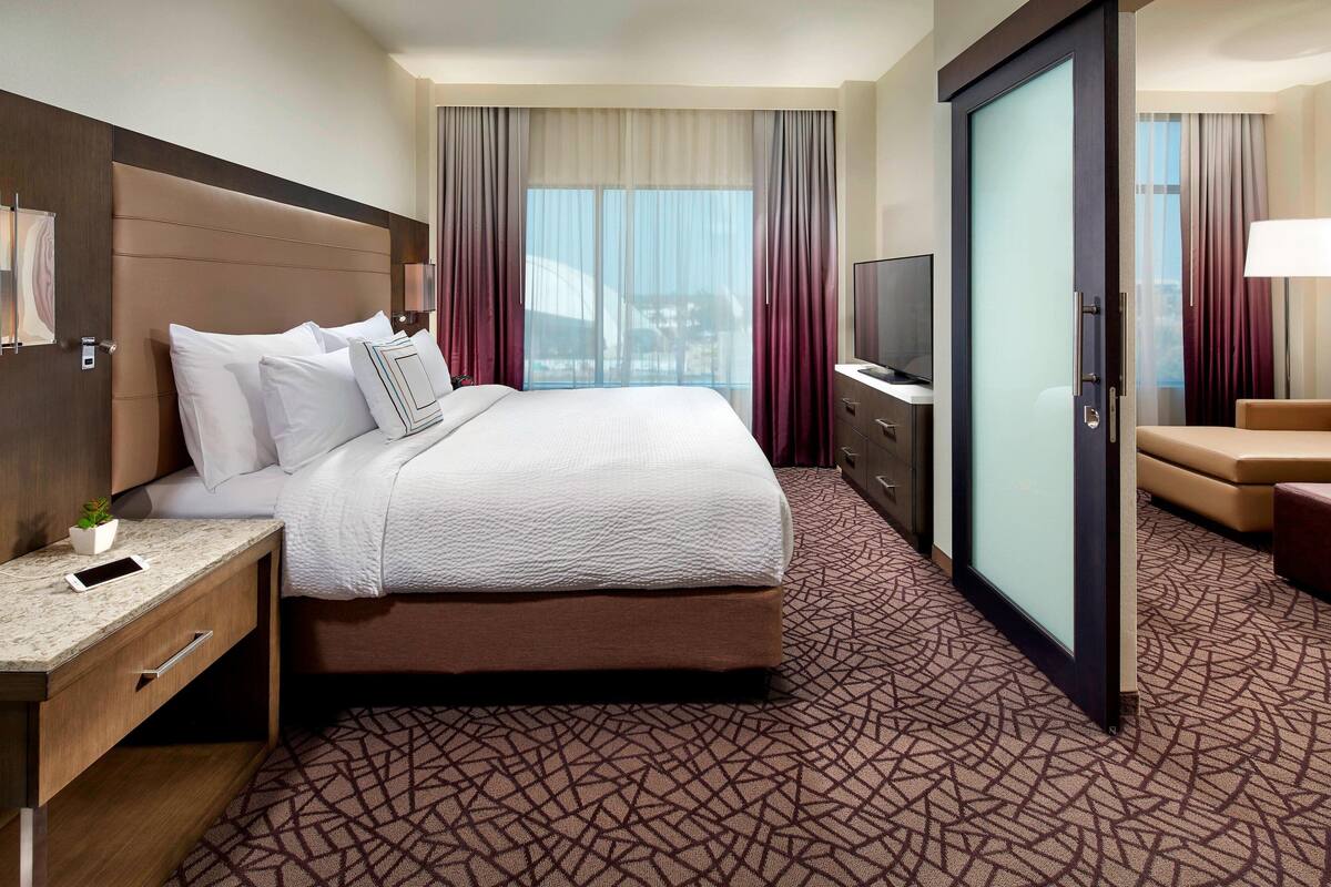 A hotel room with a large bed, a desk with a lamp, a TV, and a sliding door to a balcony.