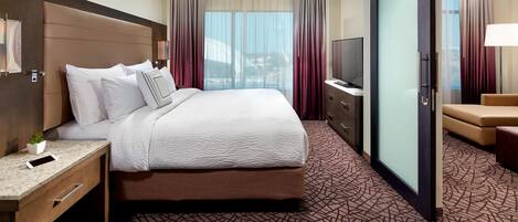 A hotel room with a large bed, a desk with a lamp, a TV, and a sliding door to a balcony.
