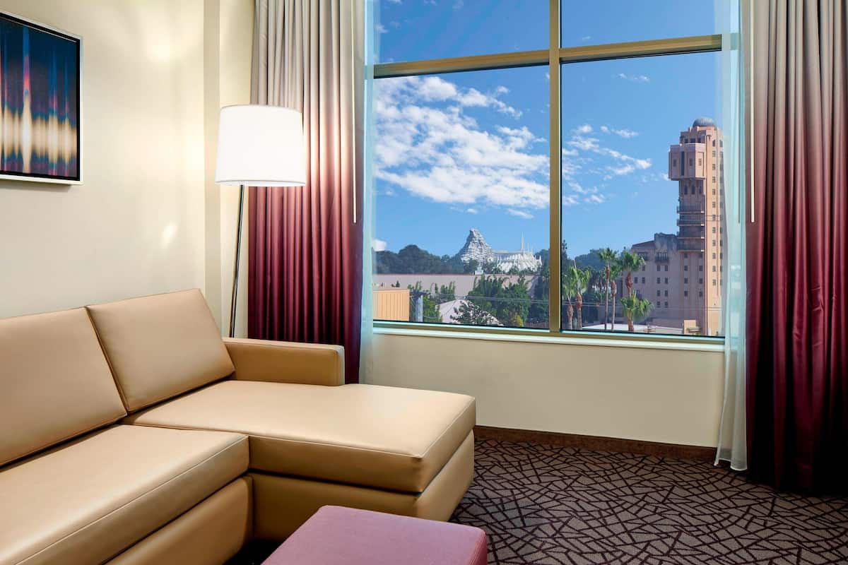 A hotel room with a beige sofa, a small pink ottoman, a floor lamp, and a large window offering a view of a cityscape and a distinctive building.