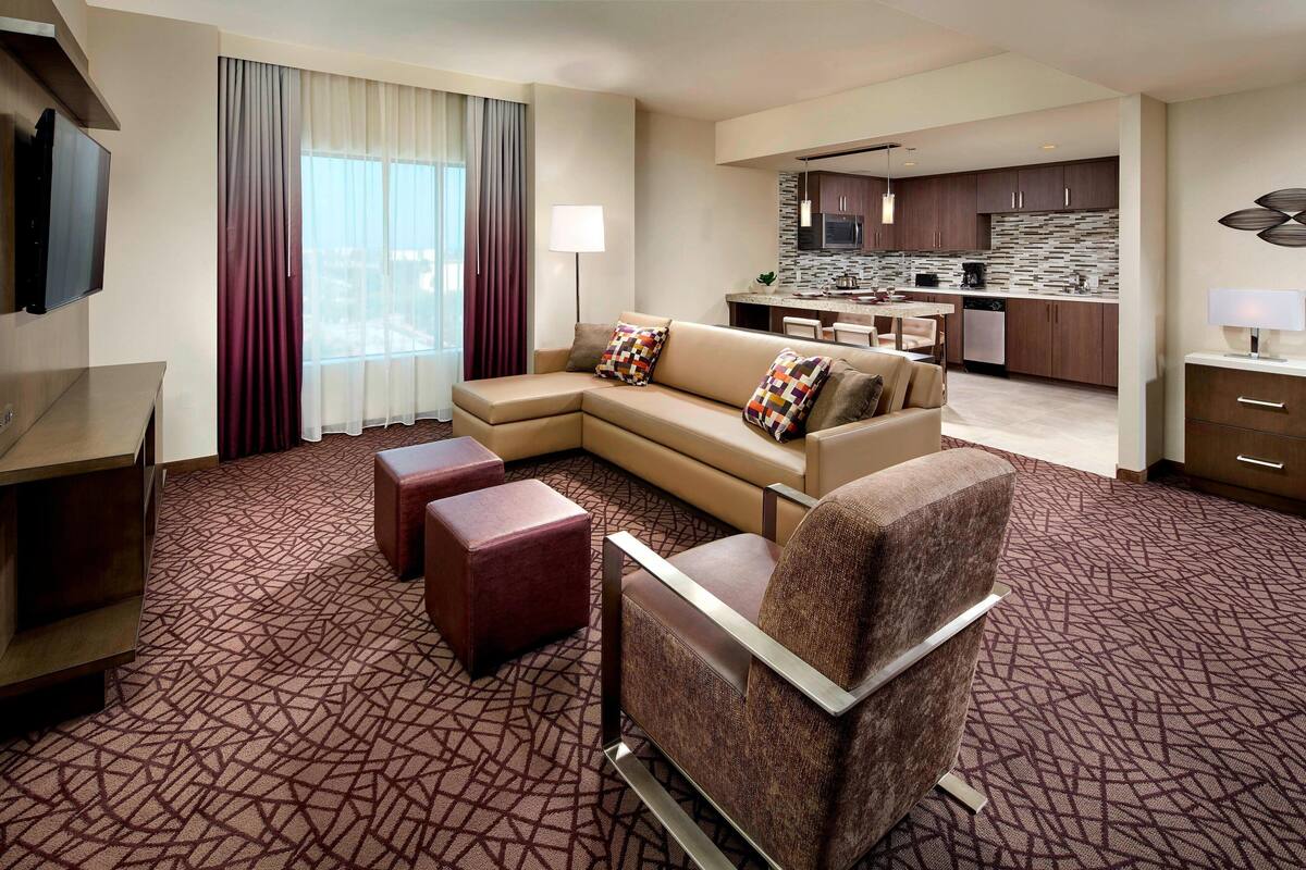 A modern hotel room with a sectional sofa, a single armchair, a small desk, and a flat-screen TV. The room features a patterned carpet and a view of the kitchen area.