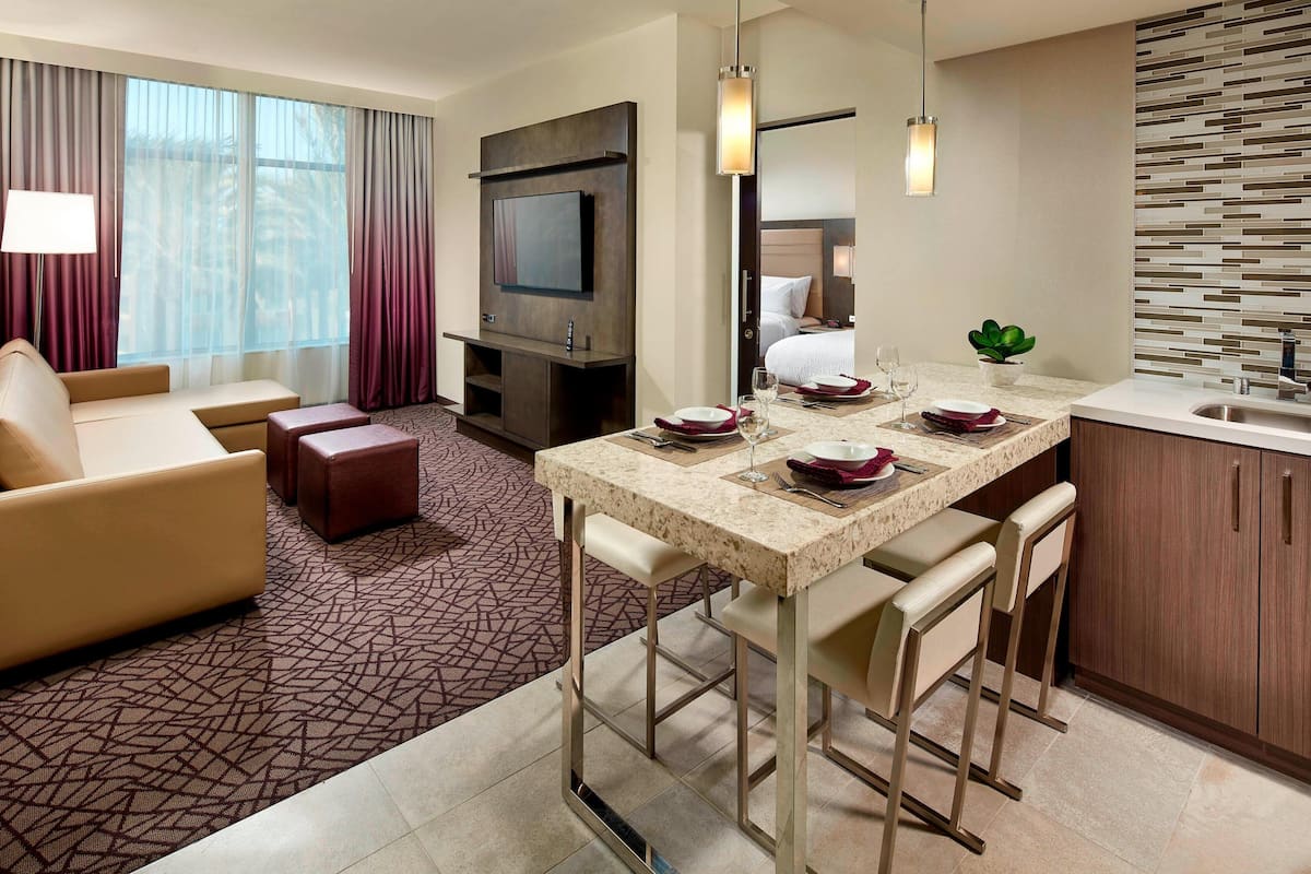 A hotel room with a sofa, a TV, a dining area with a table and chairs, a kitchen area, and a large window with curtains.
